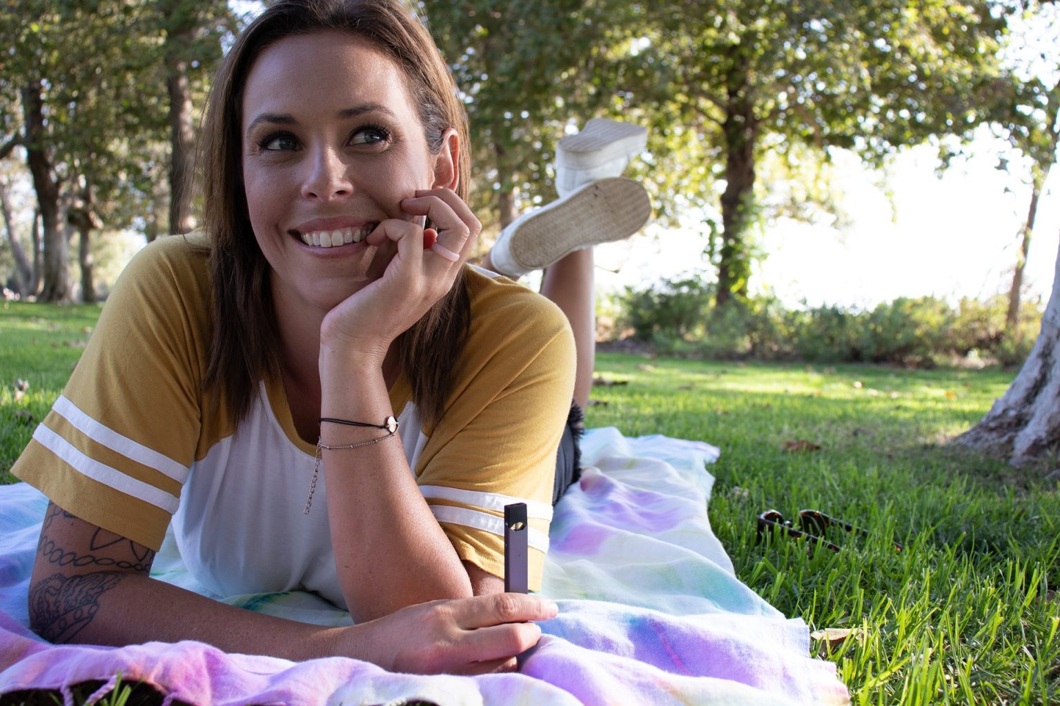 tattooed woman with brunette hair laid down and vaping a JUUL vaping device in a park