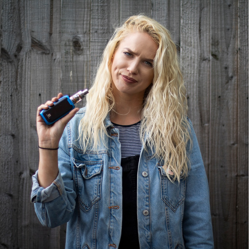 a blonde woman in blue denim jacket holding up a vaping device