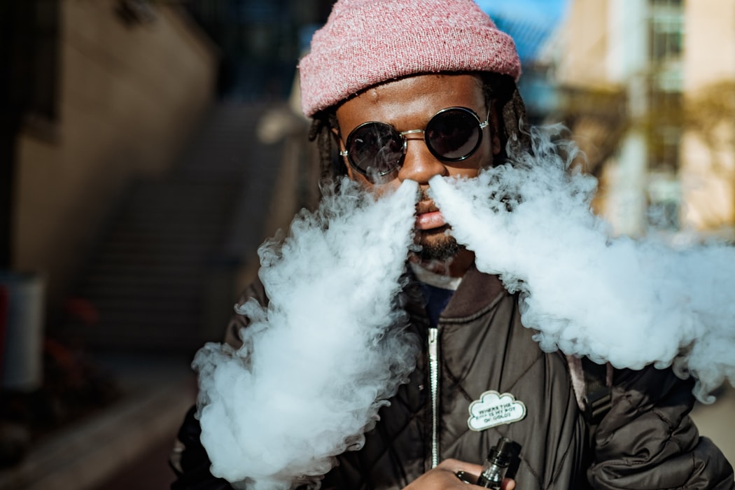 a man with a pink woolly hat and sunglasses vaping and blowing clouds through his nose with a black vaping mod