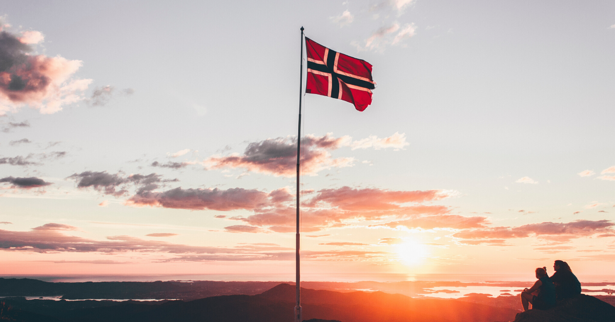 two people sat by a Norway flag on top of a mountain during sunset