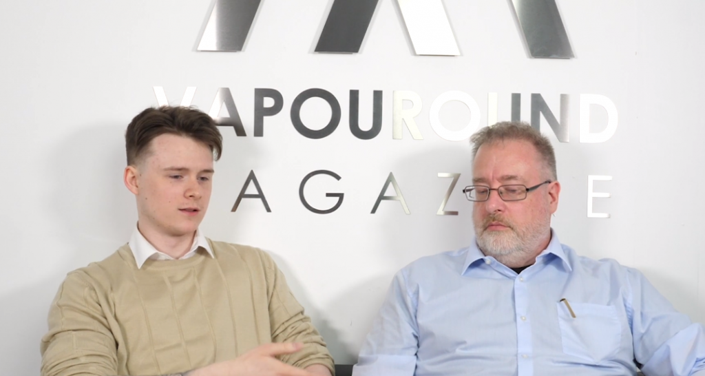 Two men sit beside each other. One is wearing a beige jumper and the other is in a blue shirt. They are in discussion in front of a sign that says Vapouround Magazine.