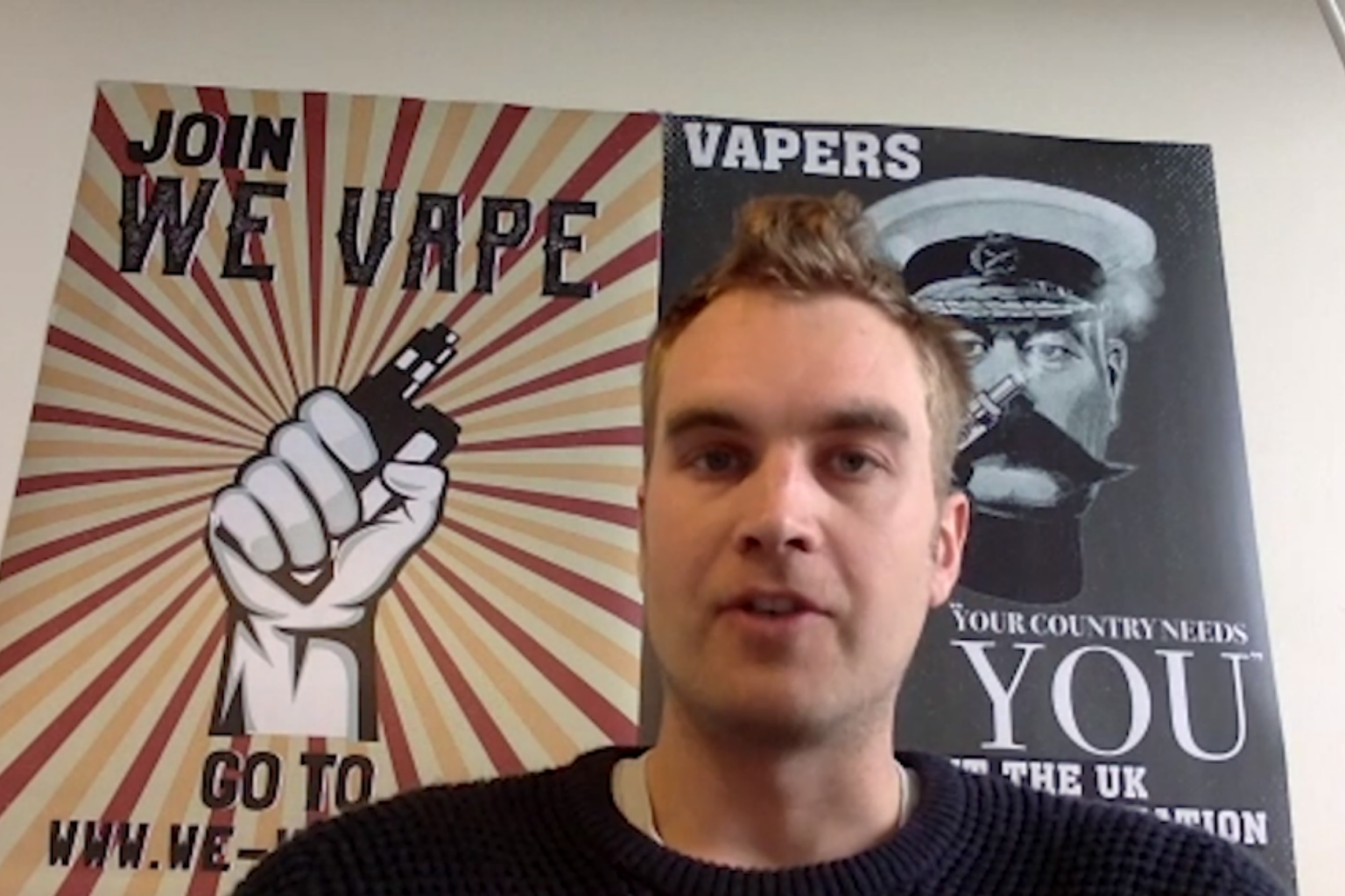 A man sits in front of a poster that says WE VAPE with a fist holding a vape device in it