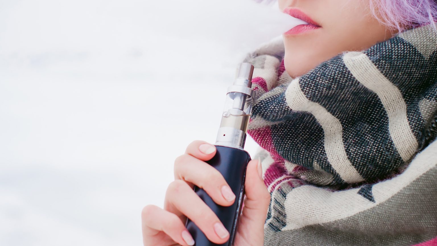 vaping girl. winter street portrait of a woman hipster, purple-dyed hair, a gray knitted hat and scarf. woman smokes an electronic cigarette in the street near the snow-covered river