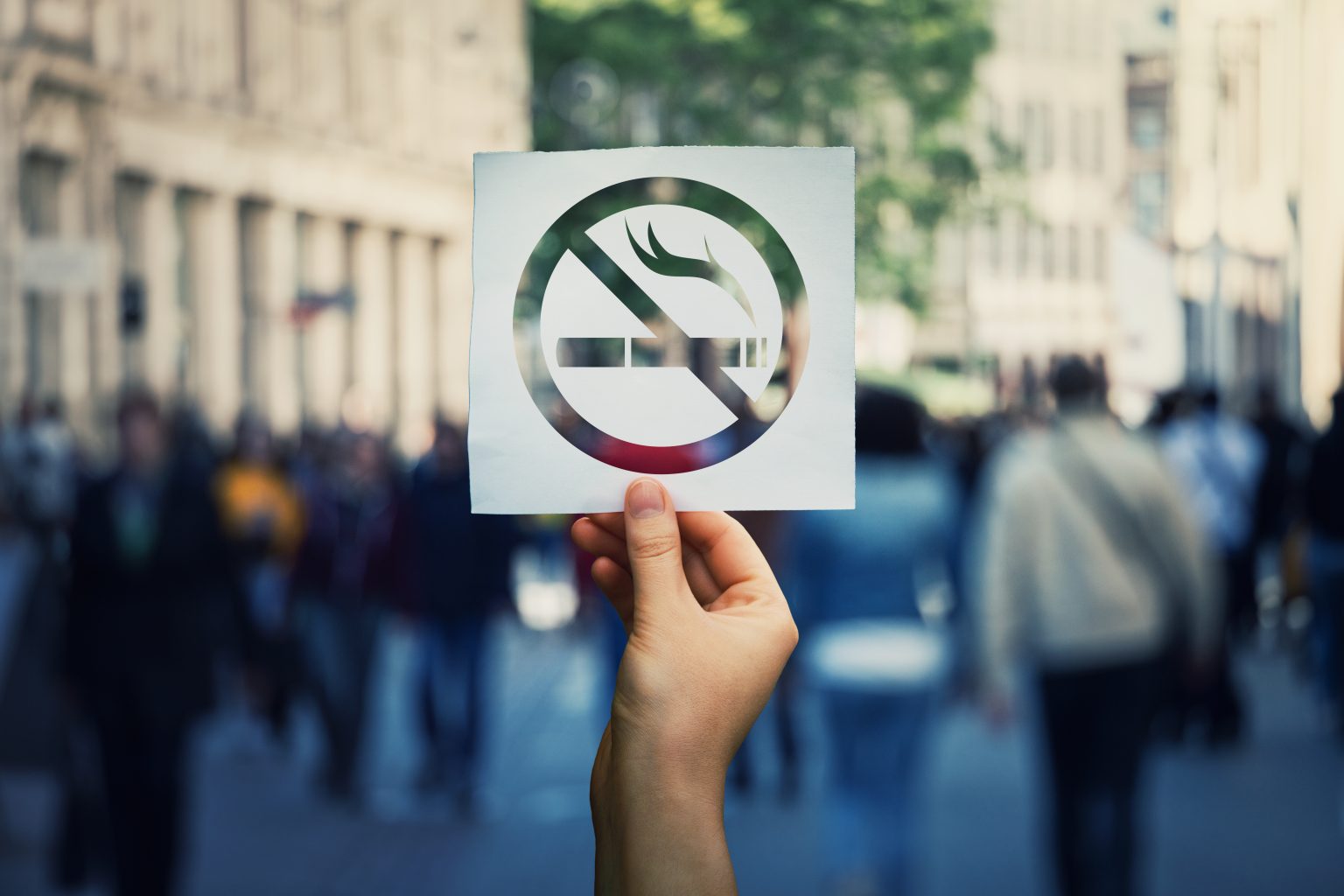 Hand holding a paper sheet with no smoking sign over a crowded street public place background. Forbidden area zone, restrictive symbol stop smoke