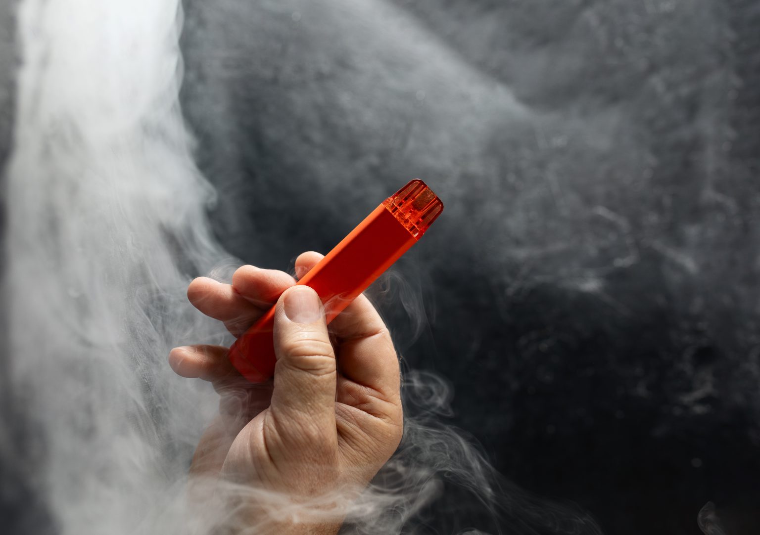 disposable electronic cigarette in a man's hand on a dark background with smoke around. The concept of modern alternative smoking, vaping and nicotine