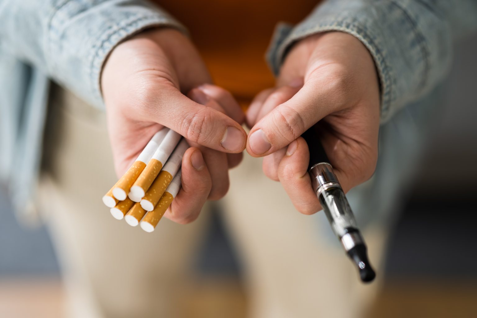 person holding tobacco cigarettes in one hand and a vape pen e-cigarette device in the other hand