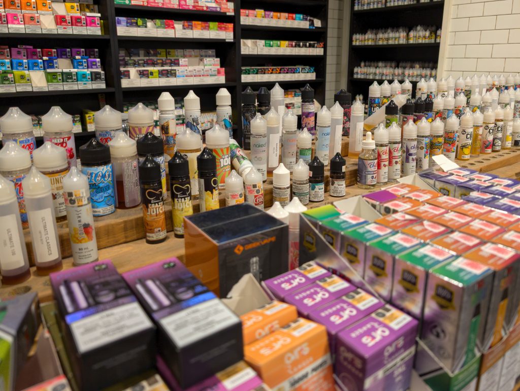 e-liquid bottles, vape juices, vape devices, disposable vapes, refillable vape kits, refillable pods displayed publicly on various shelves in a vape retail store  